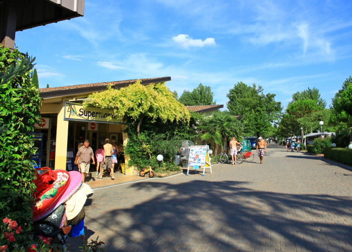 Guests walk past the supermarket at Camping Bella Italia, surrounded by trees in Veneto, Italy.