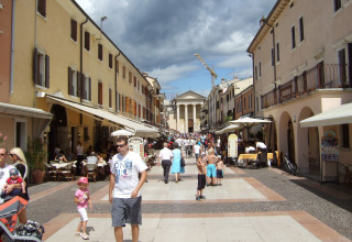 Vivace via pedonale a Camping Bella Italia, Veneto, Italia, con caffè, negozi e persone che passeggiano al sole.