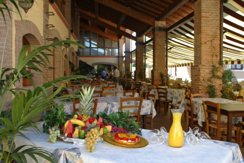 Zona de comedor cubierta en Camping Bella Italia, Veneto, con frutas frescas, zumo y mesas vacías.