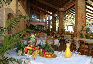 Area ristorante coperta al Camping Bella Italia, Veneto, con frutta fresca, succo e tavoli vuoti.