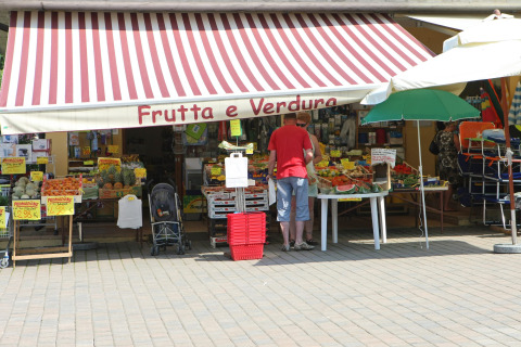 Banco di frutta e verdura a Camping Bella Italia, Veneto, Italia, con clienti sotto la tenda a strisce.