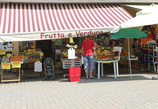 Udendørs frugt- og grøntsagsbod på Camping Bella Italia med folk, der handler under en stribet markise.