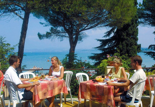 Fire mennesker nyder et udendørs måltid med udsigt over søen og træer på Camping Bella Italia i Veneto, Italien.