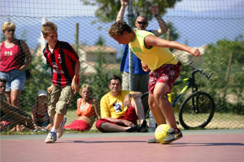 Two boys play soccer on an outdoor court at Camping Bella Italia as a crowd watches in the background.