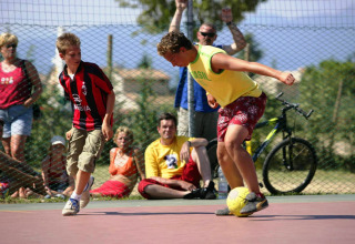 Two boys play soccer on an outdoor court at Camping Bella Italia as a crowd watches in the background.