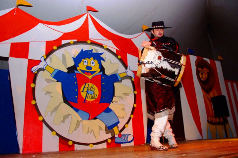 A performer in traditional outfit plays a drum on stage with circus decor at Camping Bella Italia, Italy.