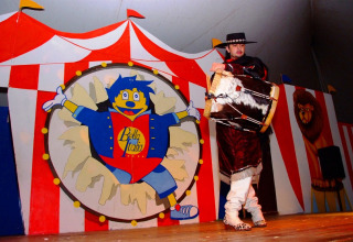 En performer i traditionelt tøj spiller tromme på en scene med farverigt cirkusdekorativ på Camping Bella Italia.