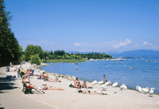 Folk solbader ved søbredden på Camping Bella Italia i Veneto, Italien, med svaner og bjerge i baggrunden.