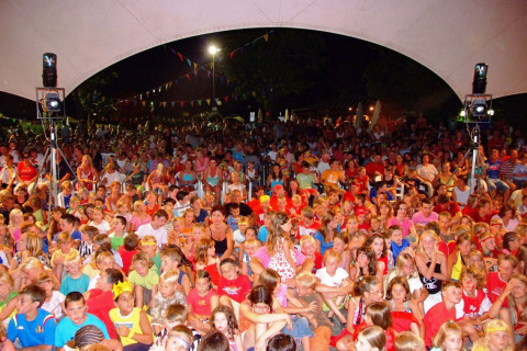 Gran grupo de niños y adultos reunidos bajo una carpa en un evento nocturno en Camping Bella Italia, Italia.