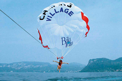 Person parasailing over a lake with a Camping Village Bella Italia parachute, mountains in the background.