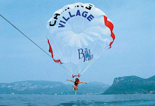 Persona fa parasailing sul lago con il paracadute Camping Village Bella Italia e montagne sullo sfondo.