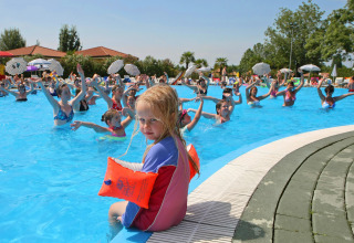 Et barn sidder ved poolkanten med baderinge, mens folk nyder svømning og aktiviteter i Camping Bella Italia.