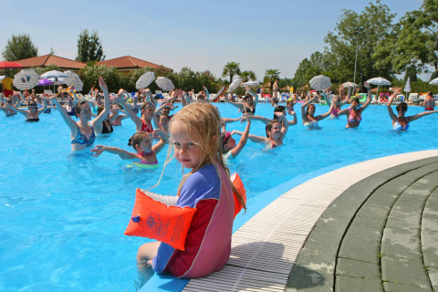Et barn sidder ved poolkanten med baderinge, mens folk nyder svømning og aktiviteter i Camping Bella Italia.