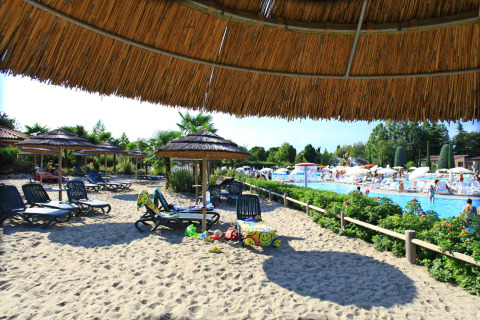 Sandy beach area with loungers and straw umbrellas by a pool at Camping Bella Italia holiday park, Veneto, Italy.