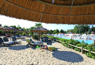 Sandy beach area with loungers and straw umbrellas by a pool at Camping Bella Italia holiday park, Veneto, Italy.