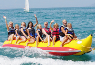 Familie nyder en tur på en gul bananbåd ved Camping Bella Italia, Veneto, Italien, med smil og bølger.