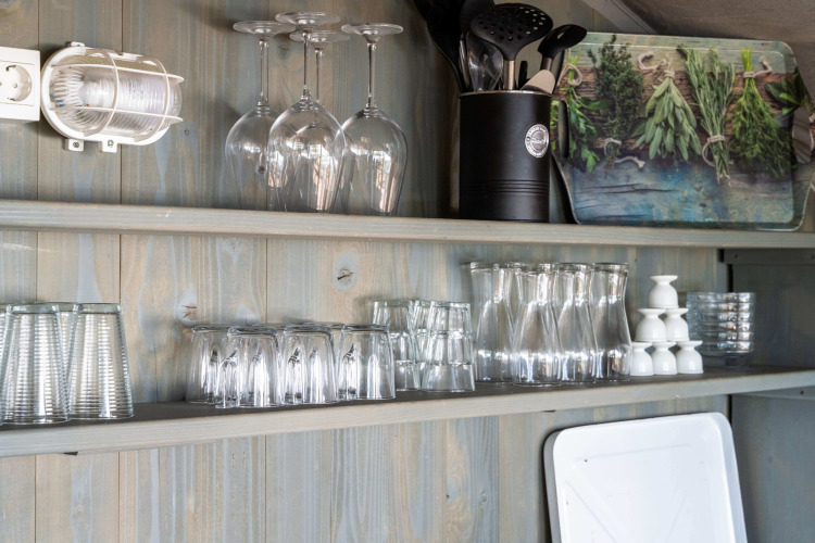 Kitchen shelves in a luxury safari tent with glasses, utensils, and a herb board on a wooden wall.
