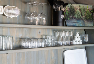 Kitchen shelves in a luxury safari tent with glasses, utensils, and a herb board on a wooden wall.