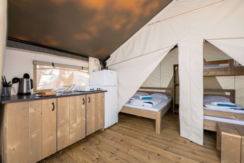 Binnenzicht van een safaritent met een keukentje, koelkast en slaapruimte met bedden en stapelbed.