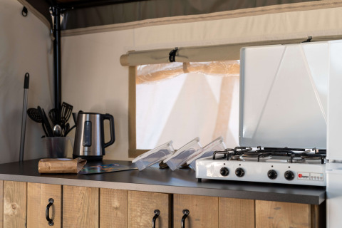 Angolo cucina in una tenda safari con bollitore elettrico, utensili da cucina e fornello a gas su banco in legno.