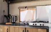 Coin cuisine d’une tente safari avec bouilloire électrique, ustensiles et gazinière sur un plan en bois.