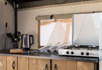 Angolo cucina in una tenda safari con bollitore elettrico, utensili da cucina e fornello a gas su banco in legno.