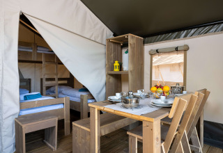 Interior of a safari tent featuring a wooden dining table, kitchenware, and wooden bunk beds in the back.
