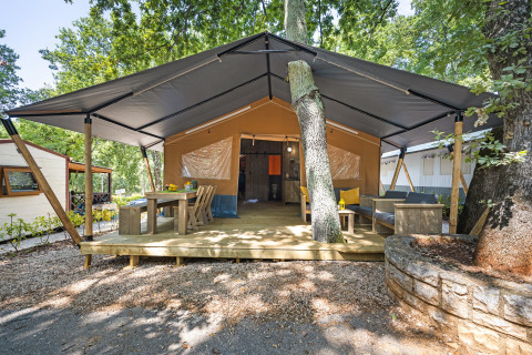 Safari tent met sanitair, houten terras, eethoek en lounge onder grote tentdoek en bomen.