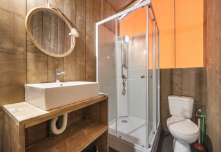 Bathroom with wooden walls, modern shower, toilet, and vanity sink inside a safari tent, cozy atmosphere.