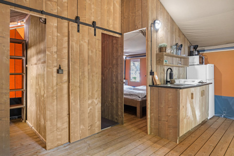 Binnenzicht van een safaritent met houten panelen, kleine keuken, slaapkamer en ladder naar de mezzanine.