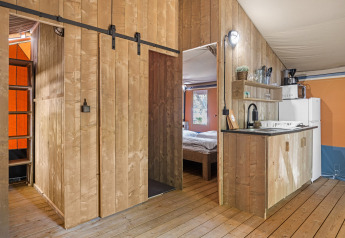 Interno di una tenda safari con pannelli in legno, angolo cottura, camera da letto e scala per il soppalco.