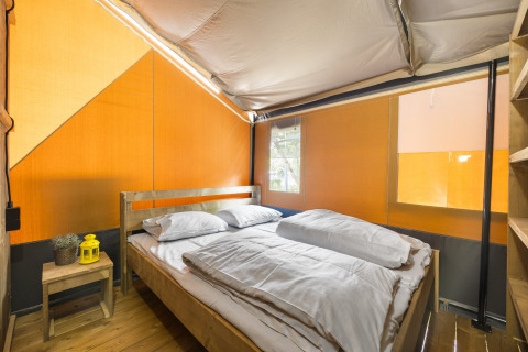Vista interior de una tienda safari con piso de madera, cama doble, ropa de cama blanca y mesa auxiliar.