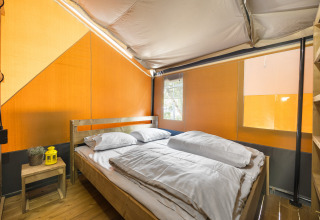 Vista interior de una tienda safari con piso de madera, cama doble, ropa de cama blanca y mesa auxiliar.