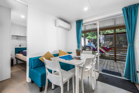 Intérieur de cabane moderne avec coin repas, rideaux bleus, table blanche et vue sur terrasse avec hamac.