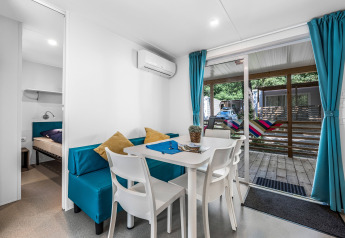 Intérieur de cabane moderne avec coin repas, rideaux bleus, table blanche et vue sur terrasse avec hamac.