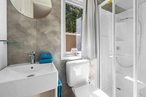 Baño moderno de cabaña con ducha, inodoro, ventana, espejo y lavabo. Toallas azules dobladas en el lavabo.