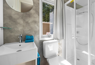 Baño moderno de cabaña con ducha, inodoro, ventana, espejo y lavabo. Toallas azules dobladas en el lavabo.