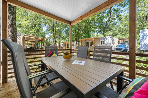 Overdekt houten terras met eettafel en stoelen, uitkijkend op moderne huisjes en omgeven door bomen.