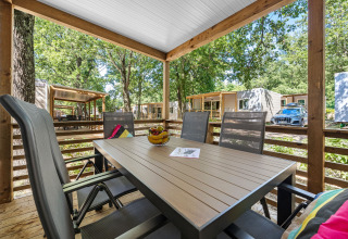 Overdækket træterrasse med spisebord og stole, omgivet af træer og moderne hytter i naturen.