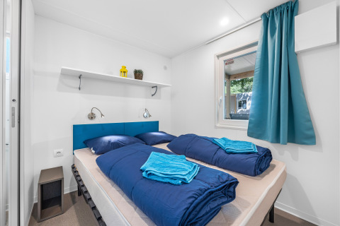 Habitación de cabina moderna con cama doble, ropa de cama azul, ventana y estante con decoraciones.