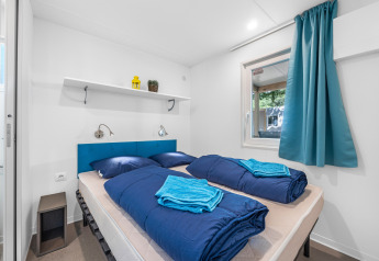 Camera moderna in cabina con letto matrimoniale, biancheria blu, finestra, tenda blu e mensola decorativa.