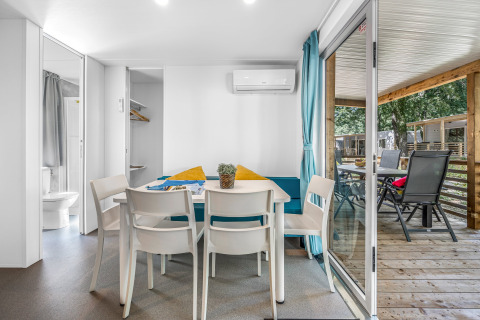 Intérieur lumineux de cabane avec table à manger, quatre chaises, vue sur terrasse couverte et mobilier extérieur.