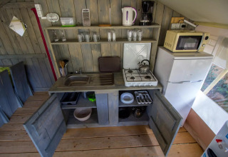 Zona cucina in una tenda safari con fornello a gas, lavandino, frigo, microonde e mobili aperti.