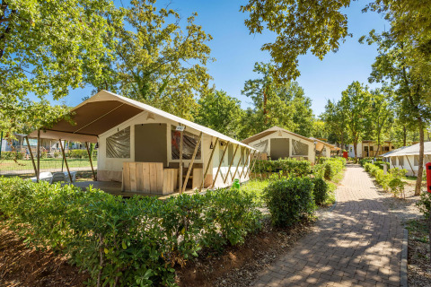 Tenda safari con servizi igienici in una zona verde, più tende lungo un sentiero pavimentato.