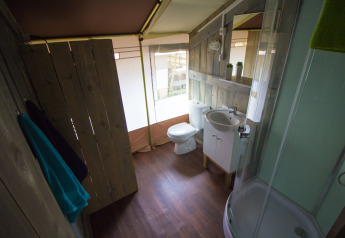 Baño en una tienda safari con suelo de madera, ducha, inodoro, lavabo, toallas y pequeñas plantas.