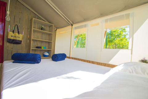 Interior de una tienda safari con cama, estantería, toallas azules y ventanas mostrando árboles verdes.