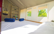 Interieur van een safaritent met bed, planken, blauwe handdoeken en ramen met uitzicht op groene bomen.