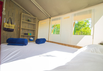 Interior de una tienda safari con cama, estantería, toallas azules y ventanas mostrando árboles verdes.