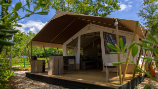 Photo d'une tente safari avec terrasse en bois et mobilier rustique, entourée d'arbres et de verdure.