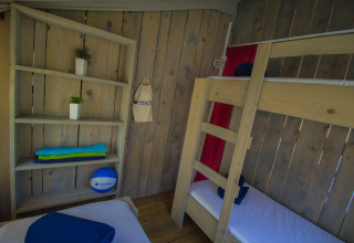 Interieur van een safaritent met stapelbedden, houten schappen en kleurrijke handdoeken op de plank.